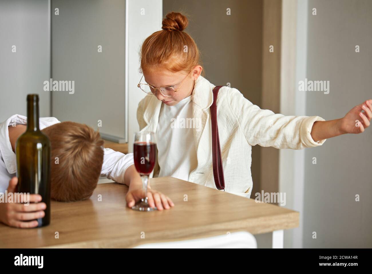 child alcoholism. little boy drinking alcohol at home, children parody adults behavior, redhead girl is next to him Stock Photo