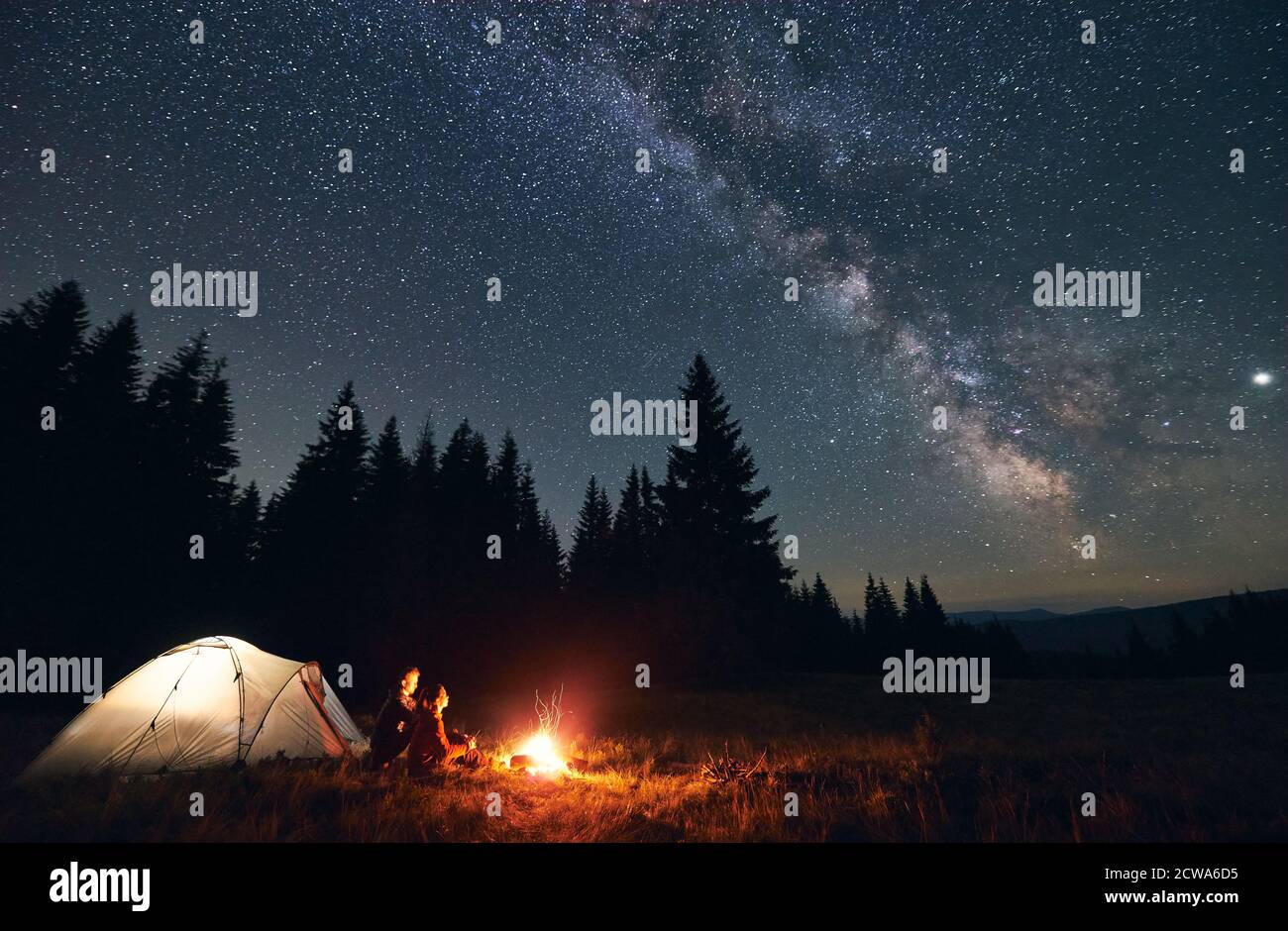 Side View Of Loving Couple Sitting Near Bright Burning Campfire And Tent Enjoying Beautiful Camping Night Together Under Dark Sky Full Of Shiny Stars And Bright Milky Way Warm Summer Night Stock