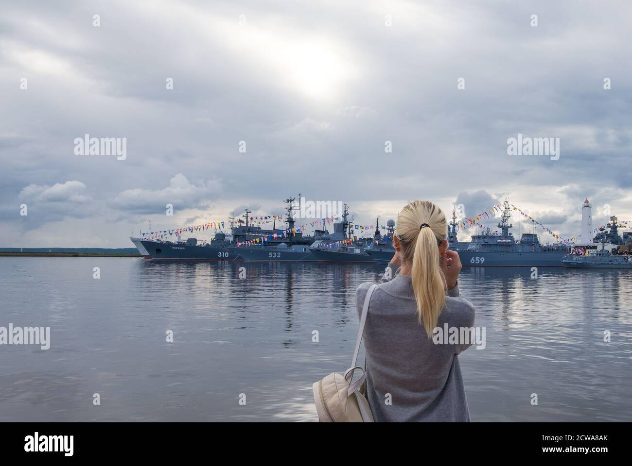 Kronshtadt, St. Petersburg: August 29, 2020. Young woman, blonde, looks at military warships with color-coded festive flags standing at the pier again Stock Photo