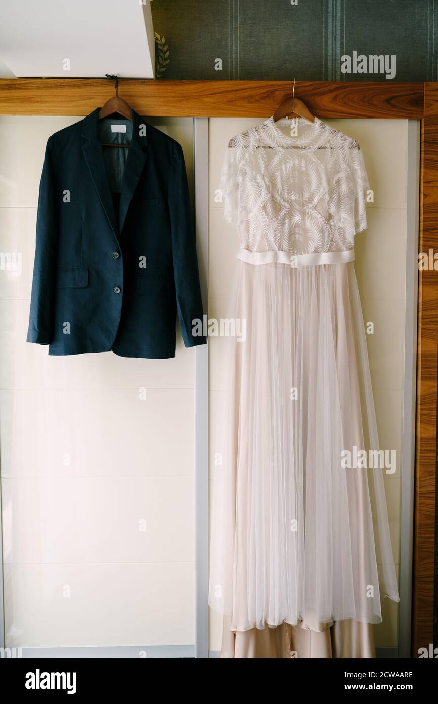 White wedding dress of the bride and jacket of the groom on wooden hangers in the room against the wall. Stock Photo