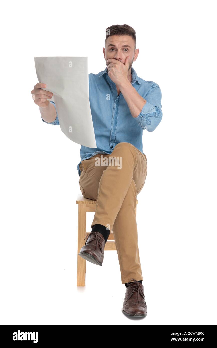 shocked young man reading newspaper and covering mouth, sitting isolated on white background, full body Stock Photo