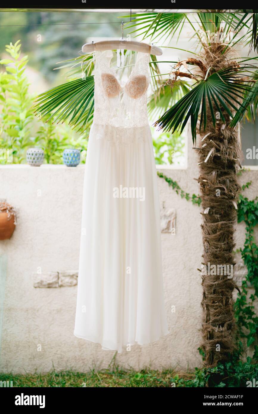 The bride's wedding dress with a lace transparent corset and a beige bodice on a hanger near a white stone fence and a decorative palm tree. Stock Photo