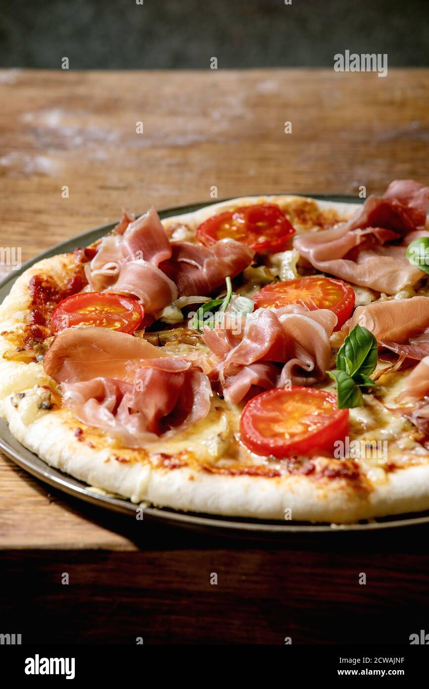 Fresh baking homemade pizza napolitana with prosciutto ham, cheese, tomatoes, basil on plate over wooden table background. Home baking or delivered fa Stock Photo