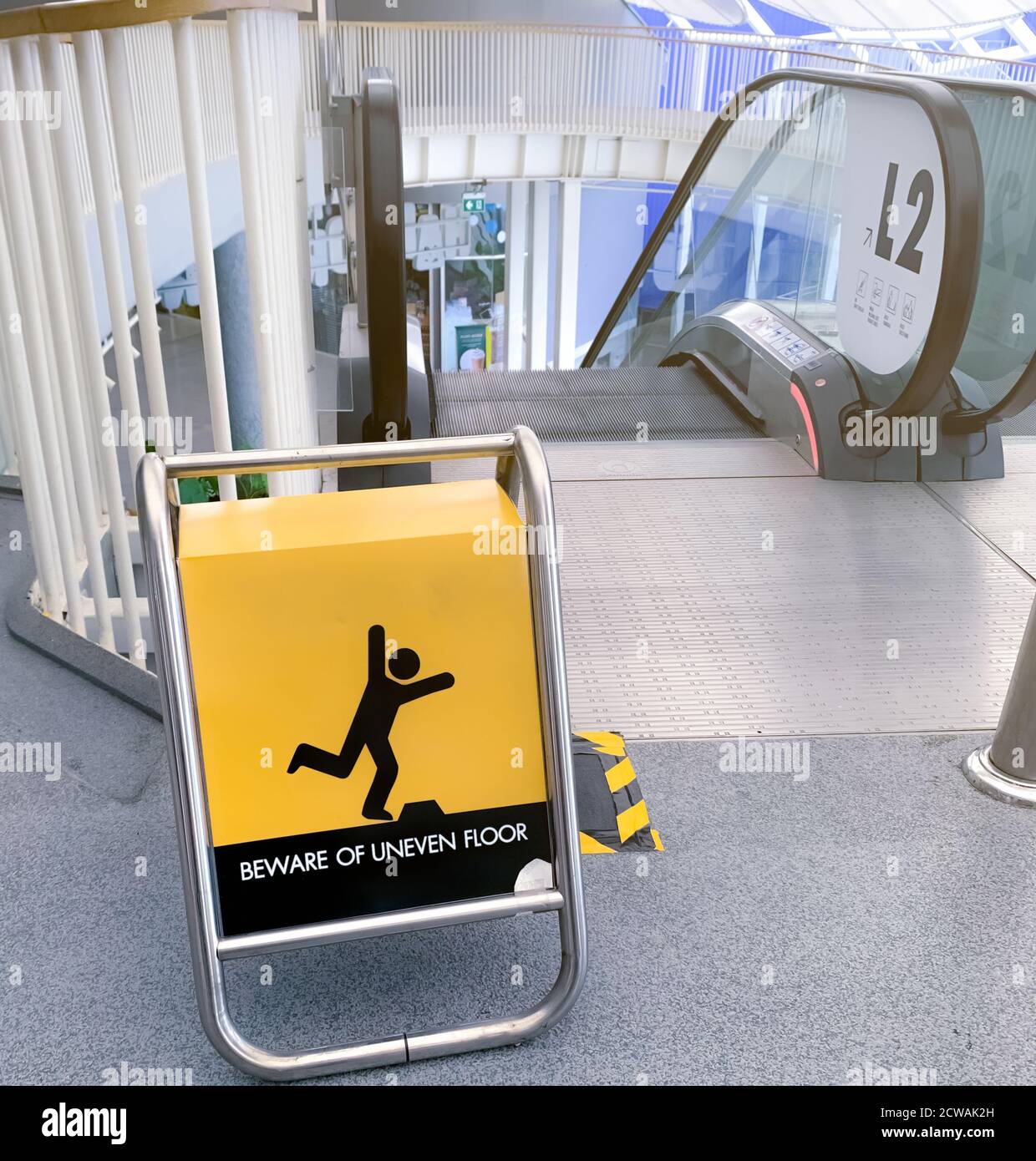 Beware uneven floor warning sign on yellow board in front of escalator in shopping mall. Beware uneven floor warning sign for safety at walkway before Stock Photo