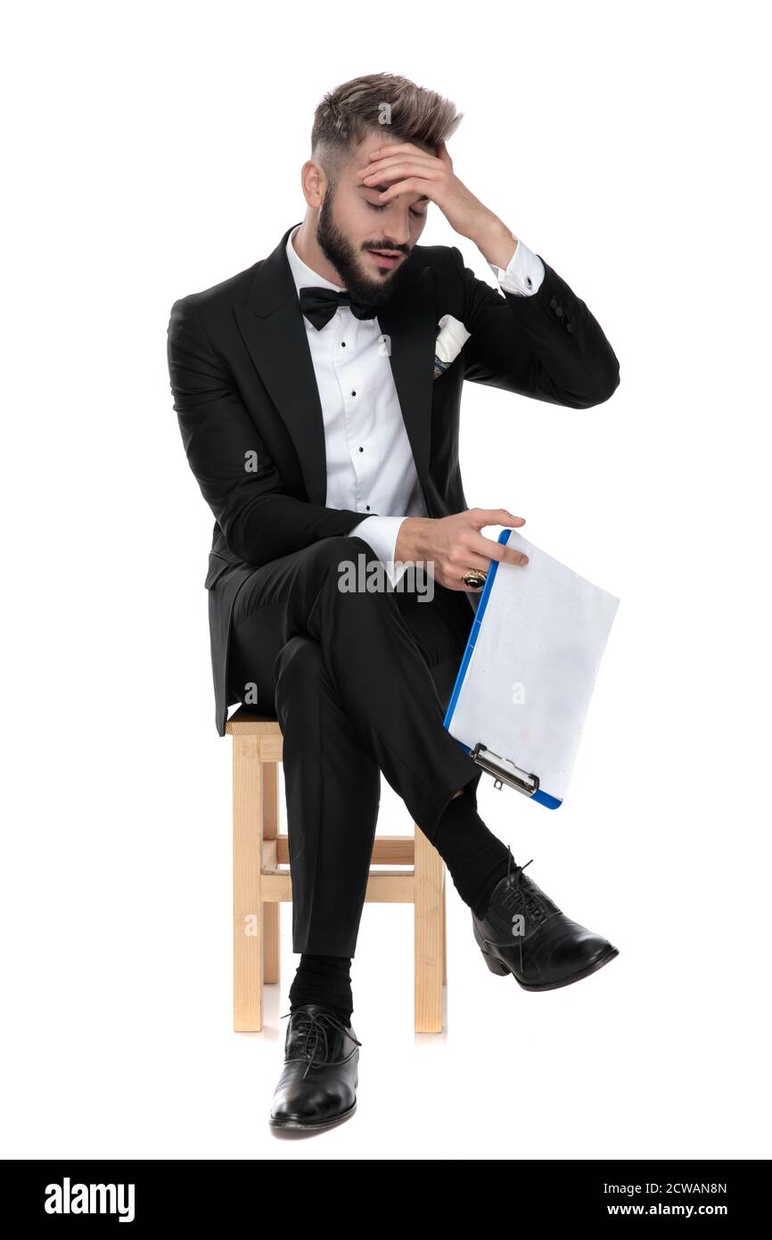 gorgeous businessman wearing black tuxedo sitting with legs crossed on a wooden chair and holding a clipboard while slapping his forehead on white stu Stock Photo