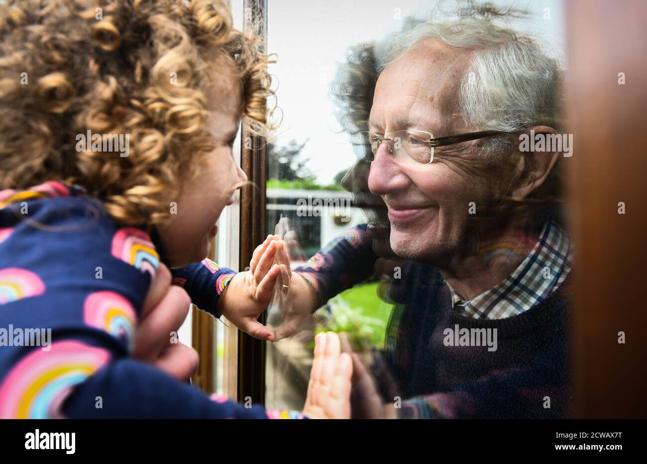 Lillian Joy aged two looks at her grandfather Edward, aged 80, through a window as meetings indoors are banned in the borough of Neath Port Talbot in Stock Photo