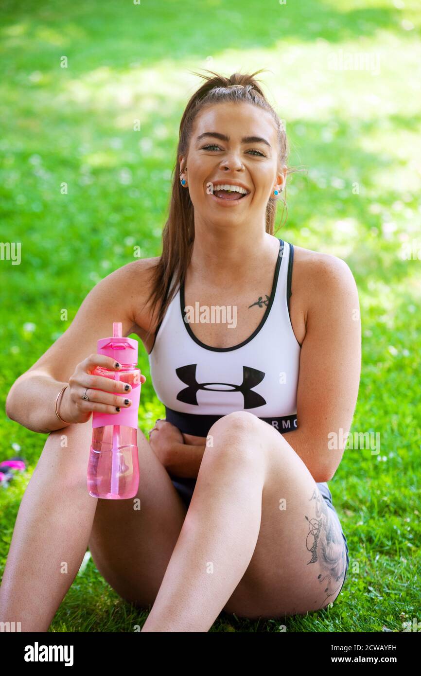 A young woman wearing sportswear sitting down outside on grass holding a pink water bottle and laughing. Stock Photo