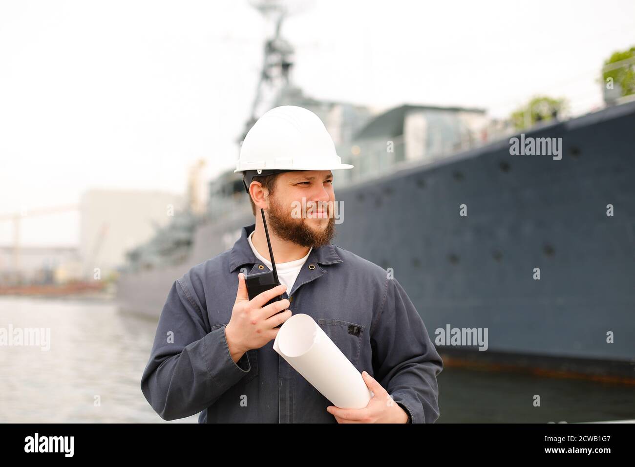 Maritime assistant engineer talking by walkie talkie, holding blueprints and standing near vessel. Stock Photo