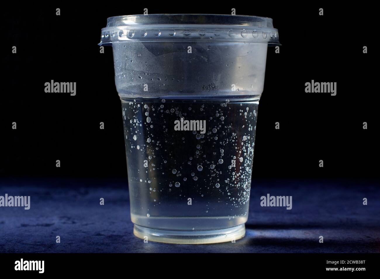 A plastic cup of fresh, cold tap water, full of air bubbles. The cup has a plastic lid on it and it is standing on deep blue background. Stock Photo