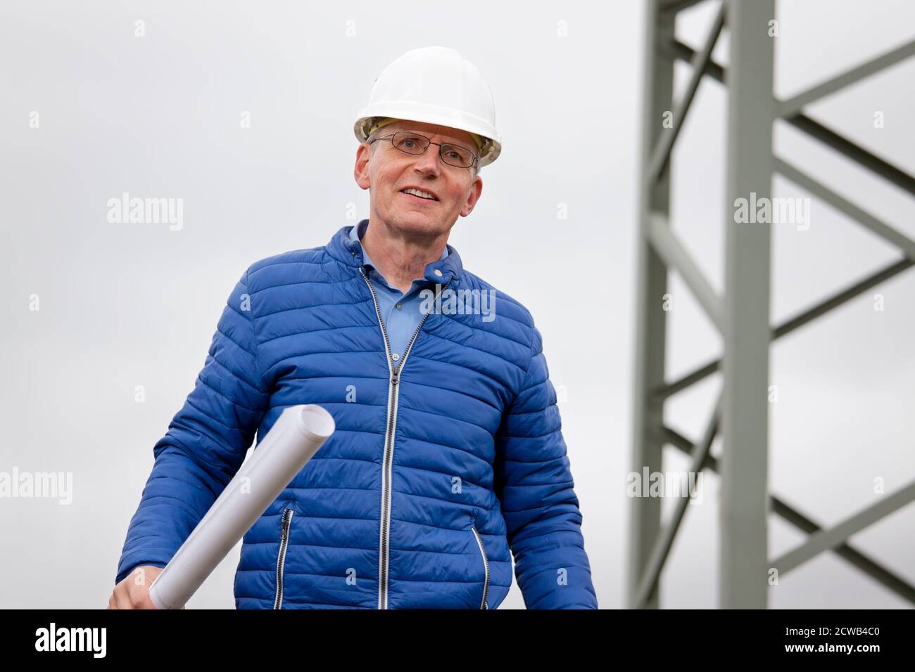 Smiling engineer with hard hat and bluepriint in his hand standing in front of an electrical pole - selective focus on the person Stock Photo