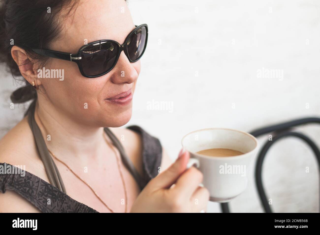 Young European woman in dark sunglasses drinks coffee with milk, outdoor portrait photo Stock Photo