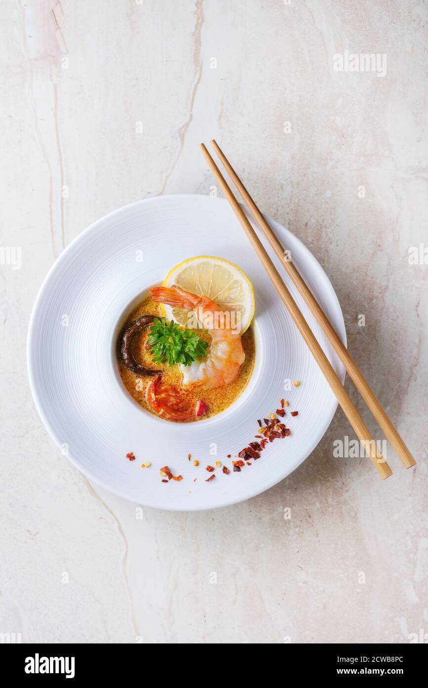 White plate with Spicy Thai soup Tom Yam with Coconut milk, Chili pepper and Shrimp over white marble as background. With wooden chopsticks. Top view. Stock Photo