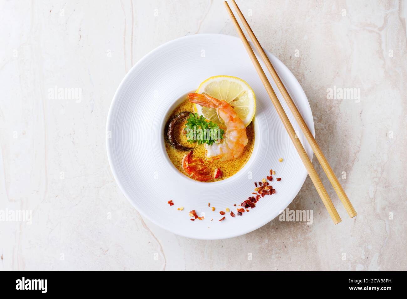 White plate with Spicy Thai soup Tom Yam with Coconut milk, Chili pepper and Shrimp over white marble as background. With wooden chopsticks. Top view. Stock Photo