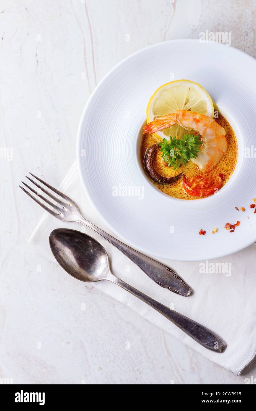 White plate with Spicy Thai soup Tom Yam with Coconut milk, Chili pepper and Shrimp over white marble as background. With fork and spoon. Top view Stock Photo