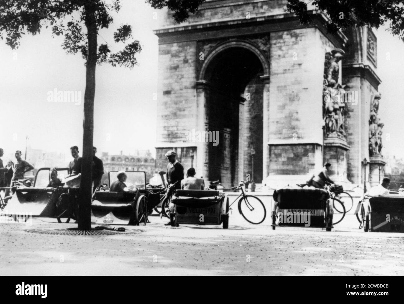 Bicycle taxis in the Place d'Etoile by the Arc de Triomphe, German-occupied Paris, August 1943. Petrol was in short supply during the occupation, with Germans using most of it. So the bicycle taxi, or taxi-velo, was a common sight on the streets of Paris. The photographer is unknown. Stock Photo
