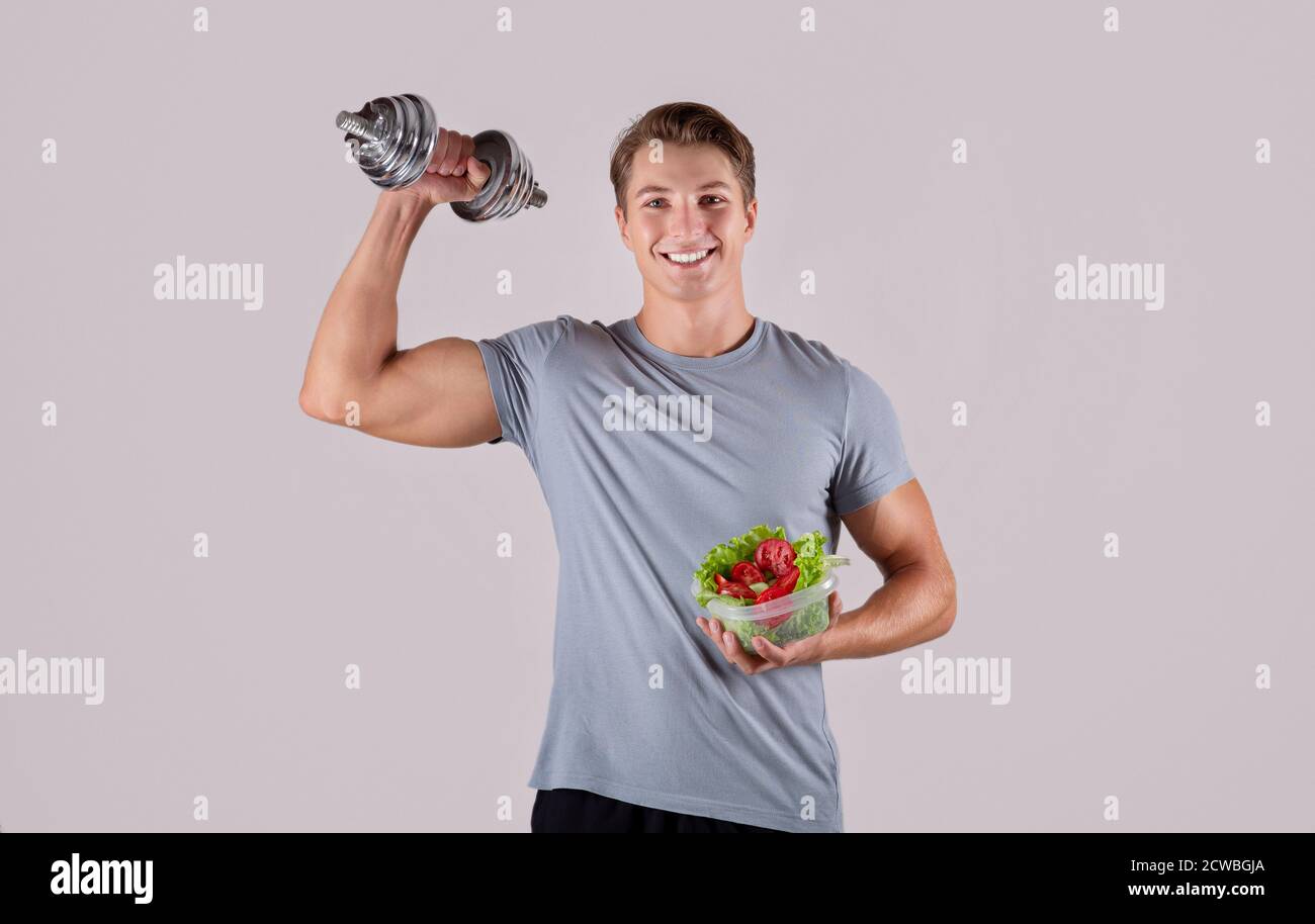 Healthy diet and exercising. Happy Caucasian guy lifting dumbbell and holding vegetable salad on light background Stock Photo