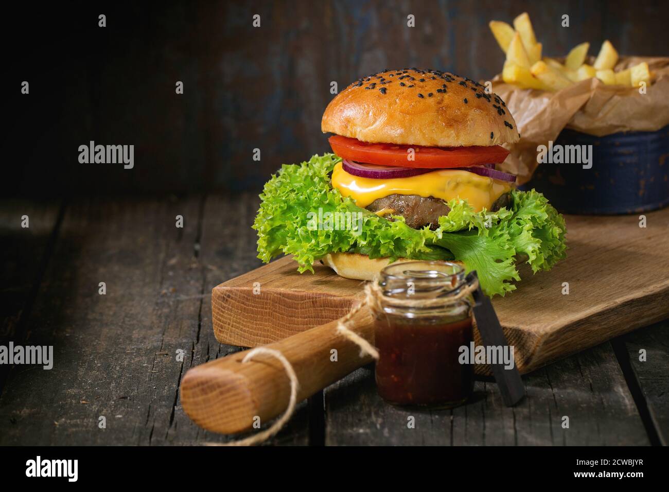 Fresh homemade burger with black sesame seeds on wooden cutting board with fried potatoes, served with ketchup sauce in glass jar over old wooden tabl Stock Photo