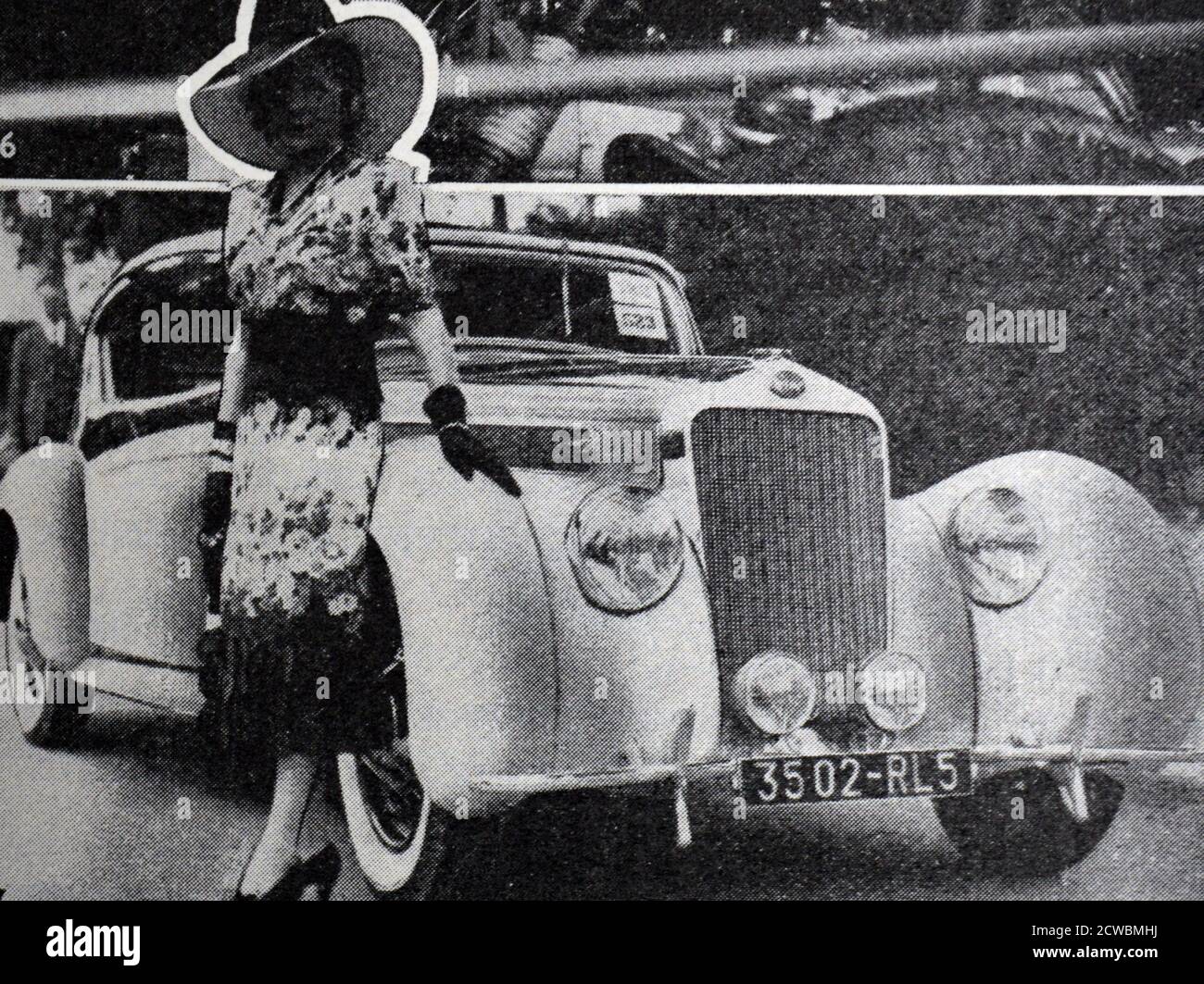 Black and white photograph pertaining to the achievements in automobile manufacturing in 1938; a competition of luxury automobiles. A well-dressed woman stands next to a new car. Stock Photo