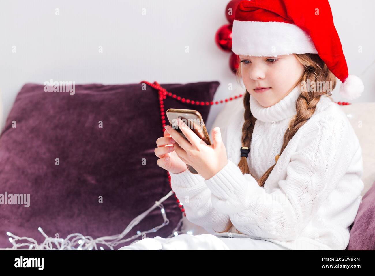 A little girl in a white sweater and a Santa Claus hat looks at a mobile phone in her hands. Stock Photo