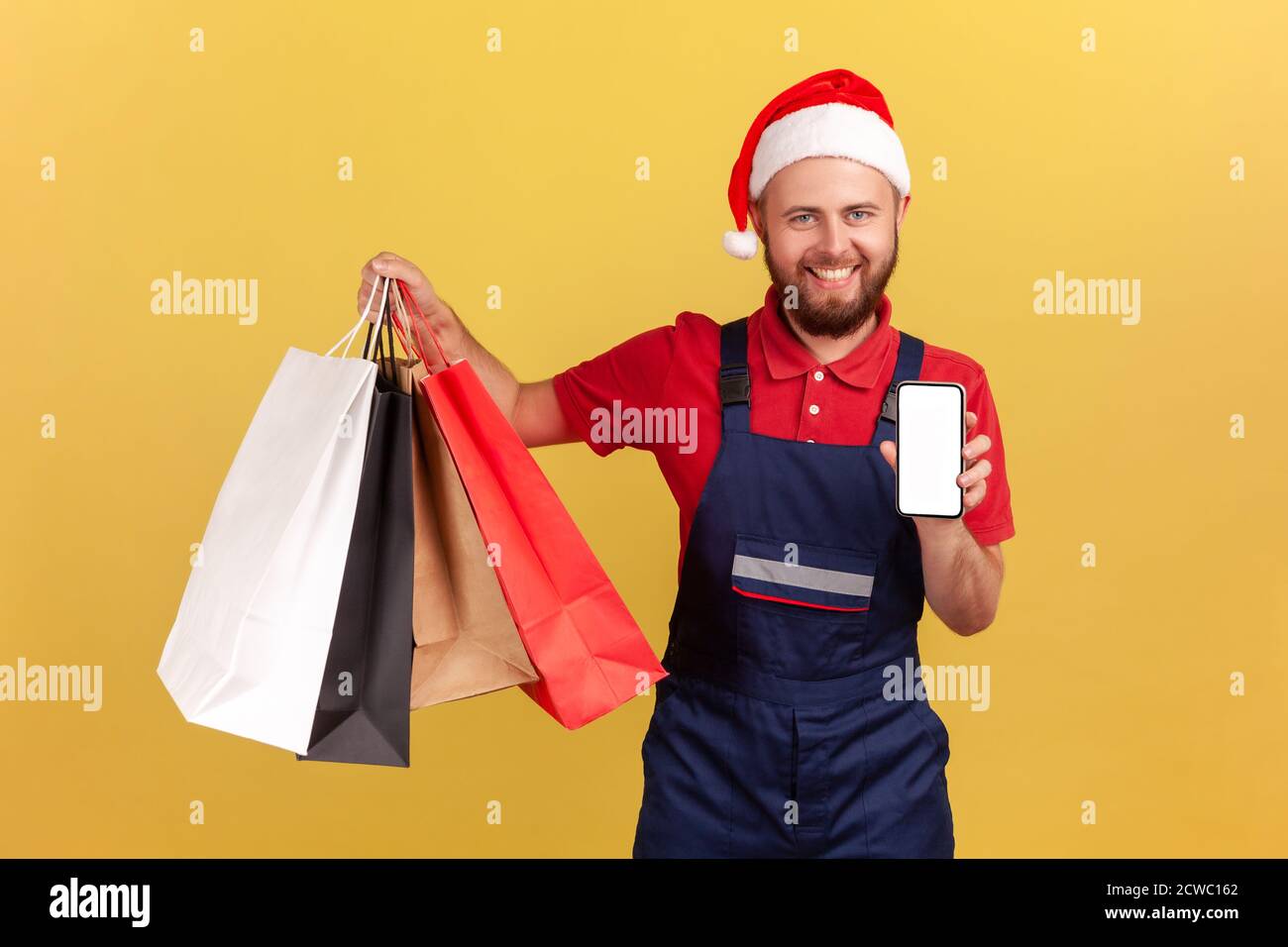 Happy positive courier in santa claus hat and blue uniform holding shopping bags and smartphone, looking at camera with toothy smile, online store. In Stock Photo