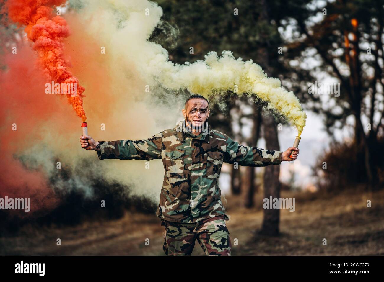 Man in camouflage uniform with black stripes on his face in the forest. In her hands, she holds red and white smoke bombs. Stock Photo