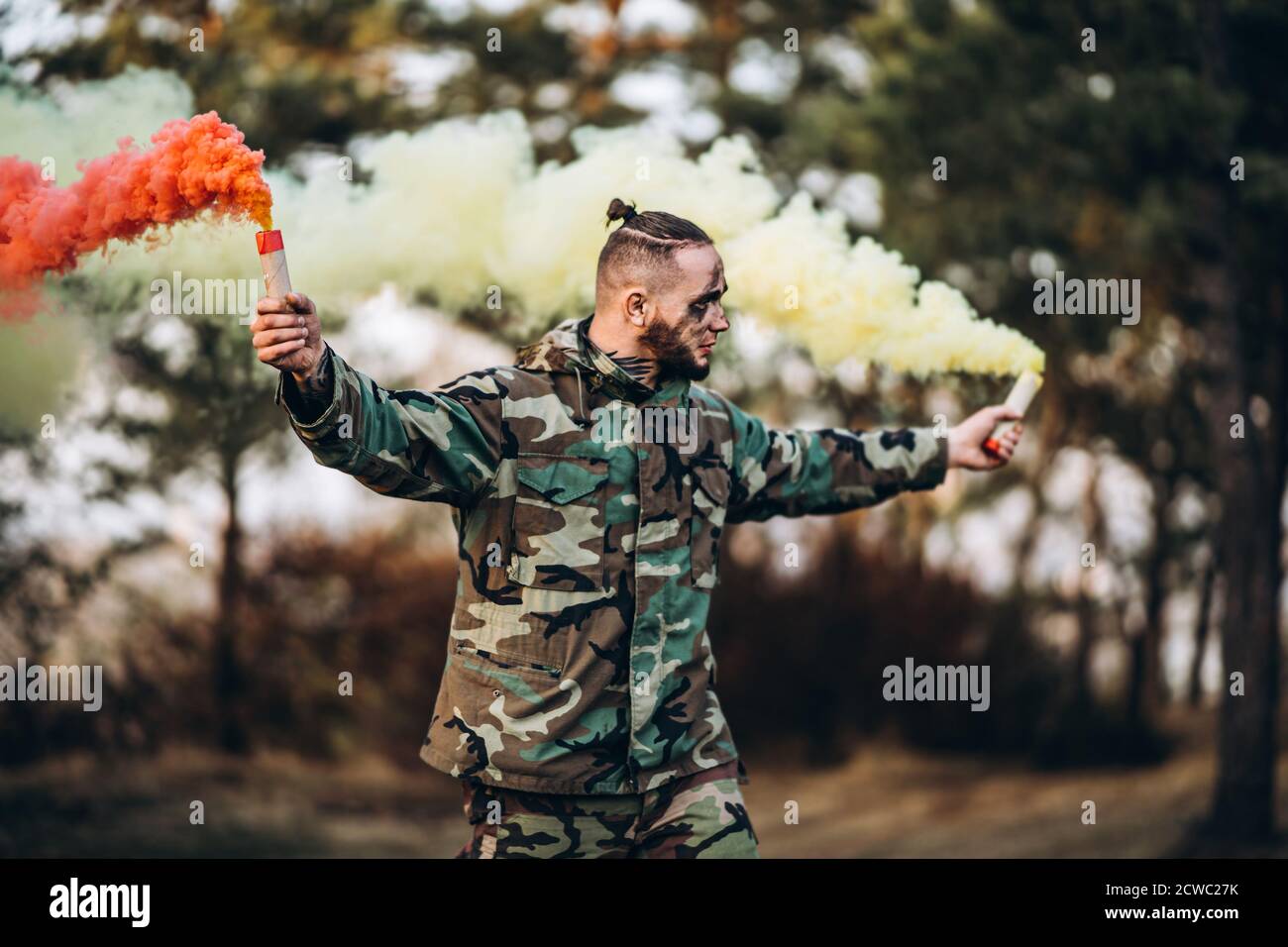 Man in camouflage uniform with black stripes on his face in the forest. In her hands, she holds red and white smoke bombs. Stock Photo