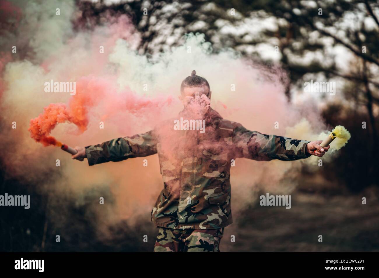 Man in camouflage uniform with black stripes on his face in the forest. In her hands, she holds red and white smoke bombs. Stock Photo