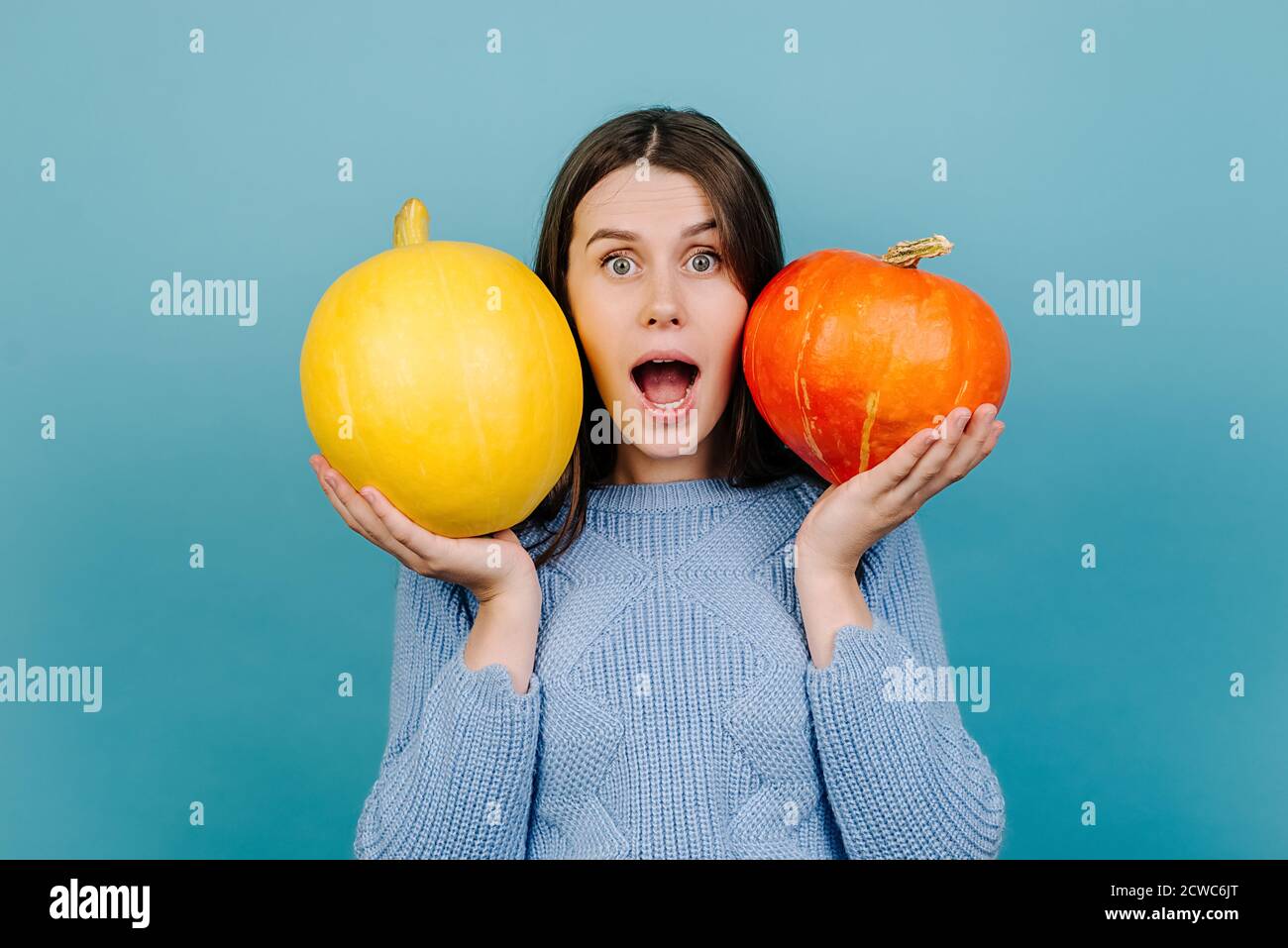 Portrait of emotional attractive young woman with open mouth, holding two small pumpkins near face, surprised looking at camera, dressed in sweater Stock Photo
