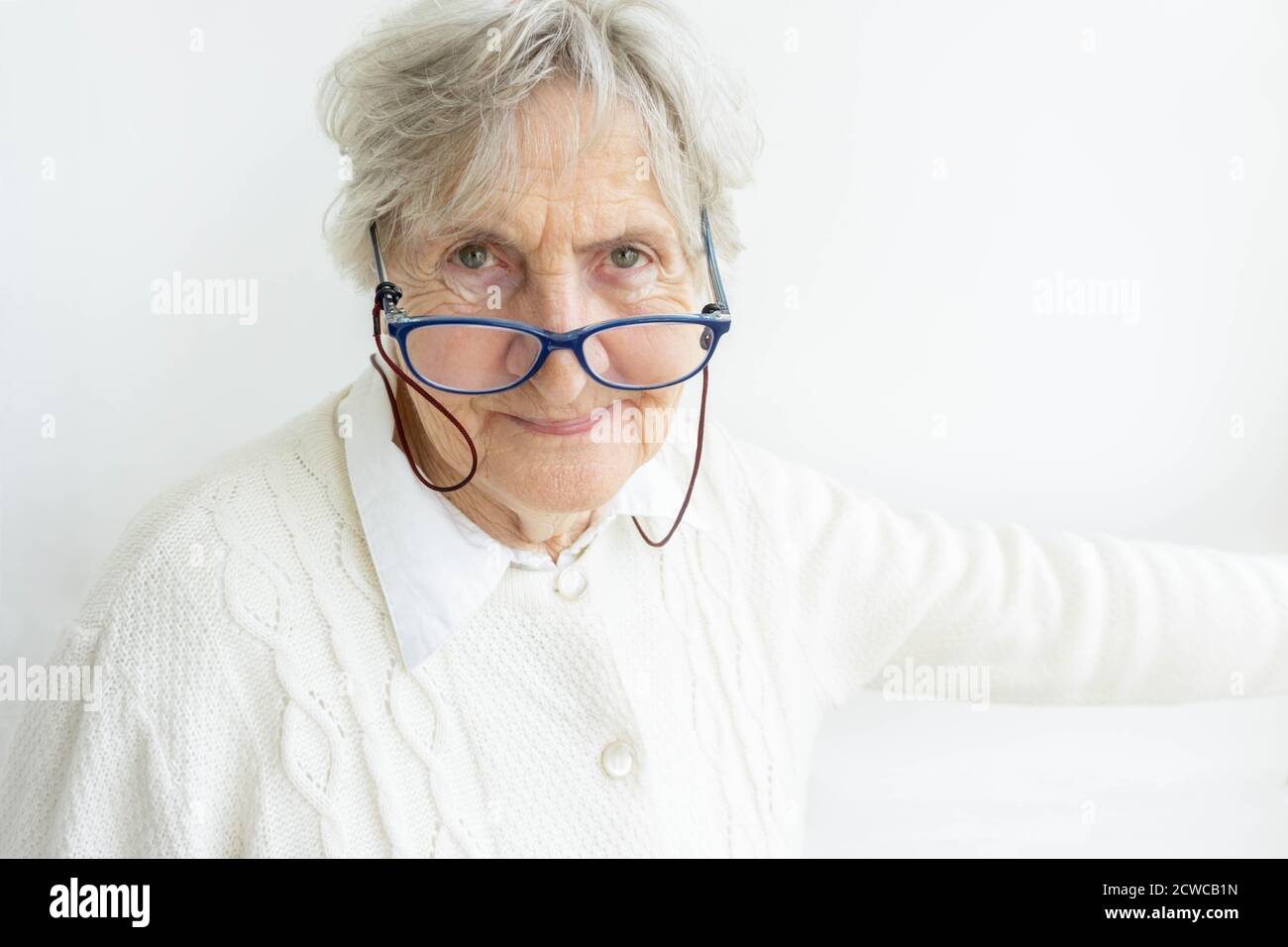 Portrait of senior woman wearing eyeglasses on wall background. Old woman in white sweater looking at camera. Gray hair senior lady in glasses isolate Stock Photo