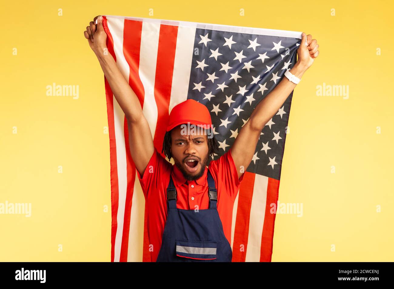 Serious afro-american worker in uniform with dreadlocks holding American flag and screaming, celebrating freedom and independence. Indoor studio shot Stock Photo