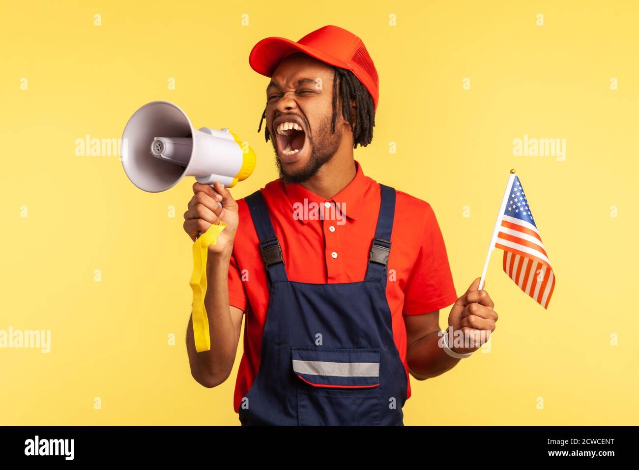 Angry nervous afro-american worker in uniform with beard and dreadlocks holding usa flag screaming at loudspeaker, protesting. Indoor studio shot isol Stock Photo