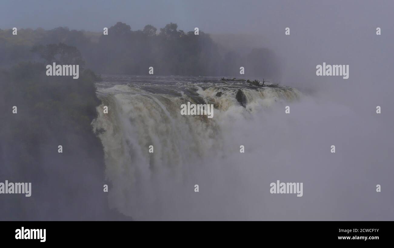 Impressive view of Zambezi River rushing down Victoria Falls seen through enormous water spray with rainforest near the border of Zimbabwe and Zambia. Stock Photo