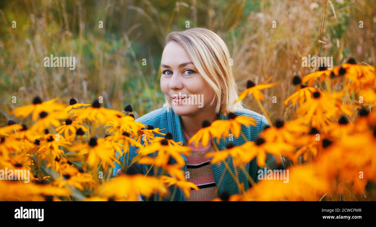 Smiling young woman sitting in the flowers. Blonde female autumn outdoor portrait with blooming flower meadow Stock Photo