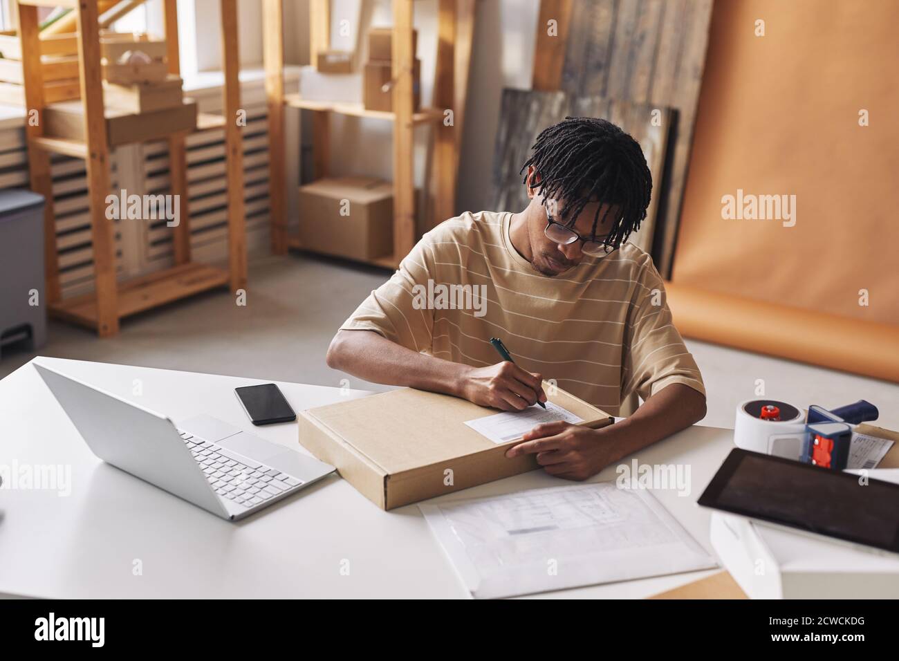 African young man sitting at the table in front of the laptop and writing address on the box before shipment Stock Photo