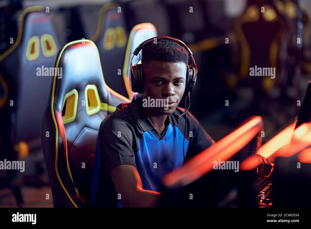 Portrait of a happy african guy, professional gamer wearing headphones looking at camera and smiling while participating in eSport tournament, sitting in gaming club or internet cafe Stock Photo