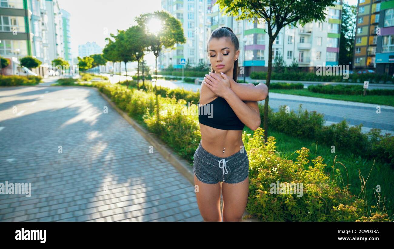 Muscular young woman stretching arms before workout near multistorey buildings in sunny morning. Girl practicing relaxing exercises, warming up upper body outdoors. Sport, flexibility concept. Stock Photo