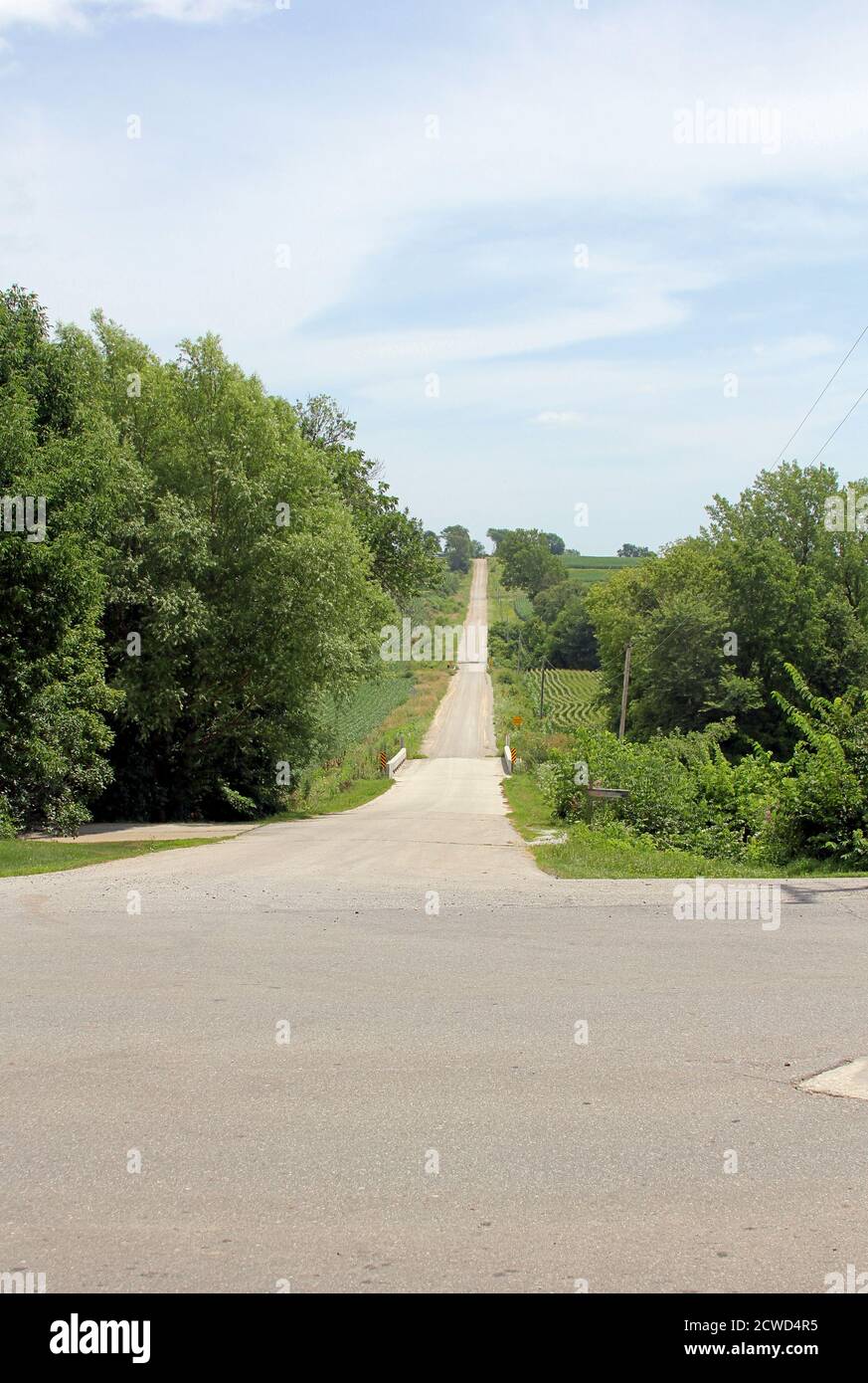 Country road leading through the fields and trees, over a bridge and up a hill into the distance. Stock Photo