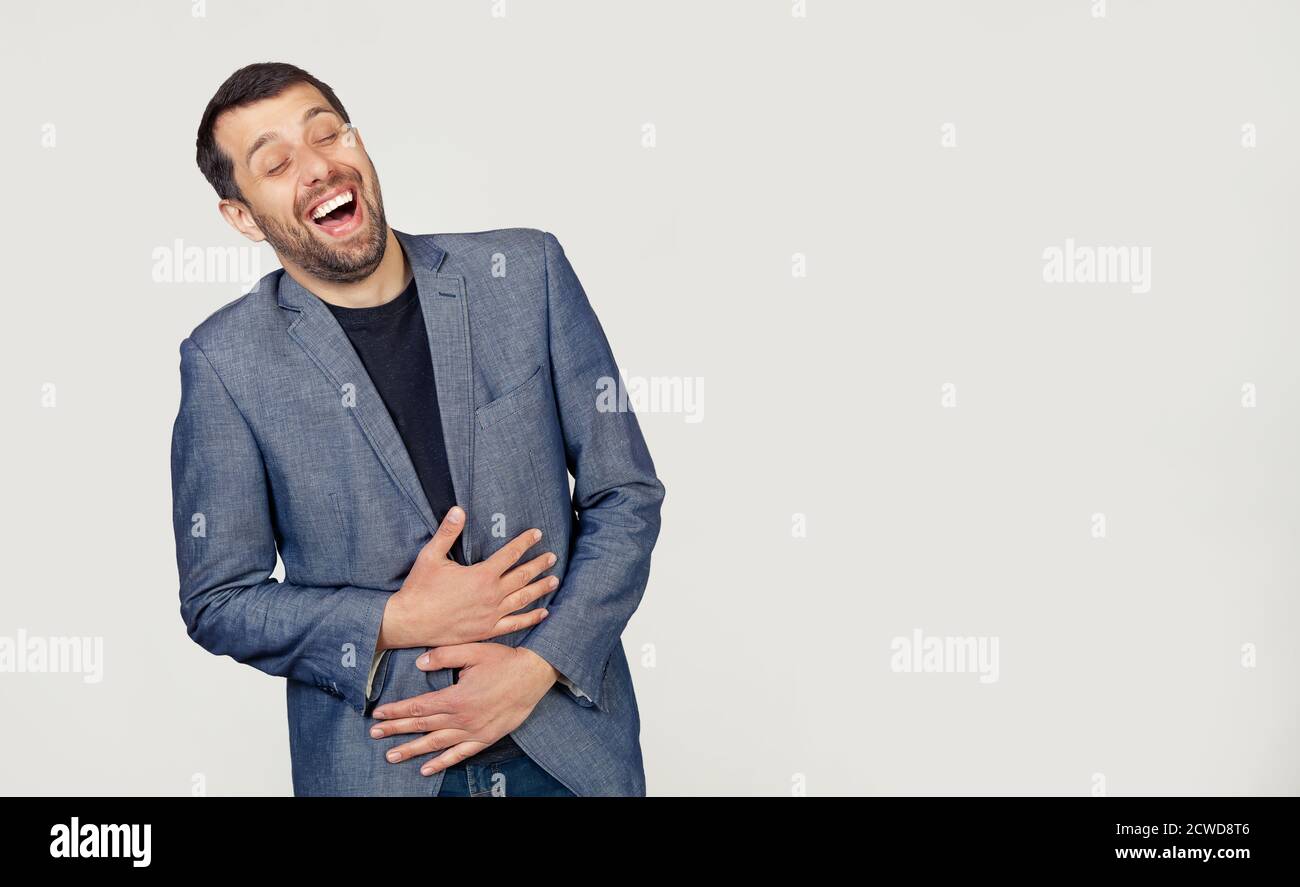 Young businessman man with a beard in a jacket smiling and laughing out loud because a funny crazy joke with hands on the body. Portrait of a man on a gray background. Stock Photo
