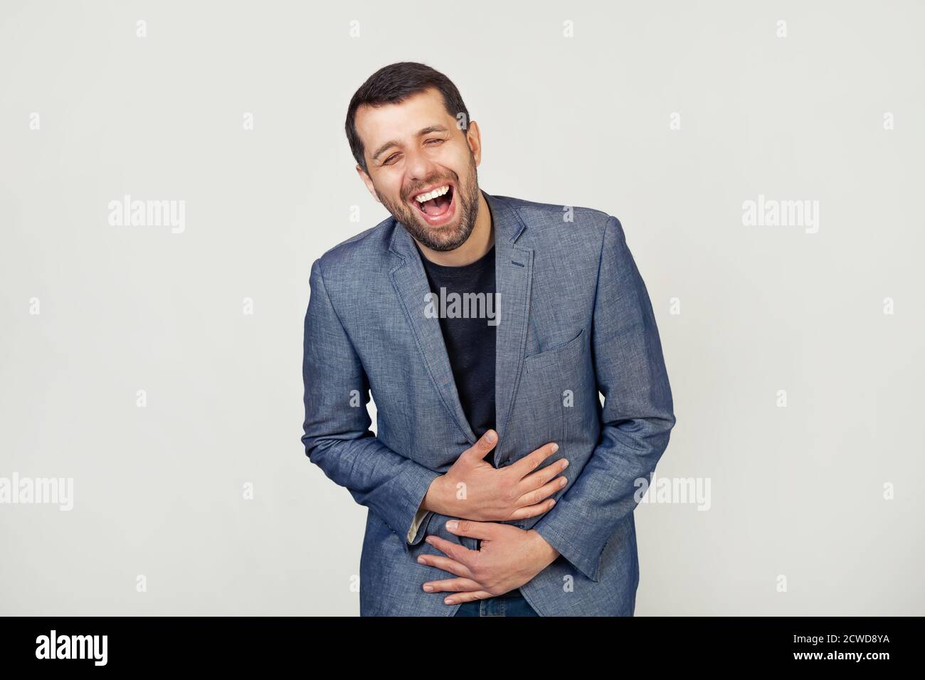 Young businessman man with a beard in a jacket smiling and laughing out loud because a funny crazy joke with hands on the body. Portrait of a man on a gray background. Stock Photo