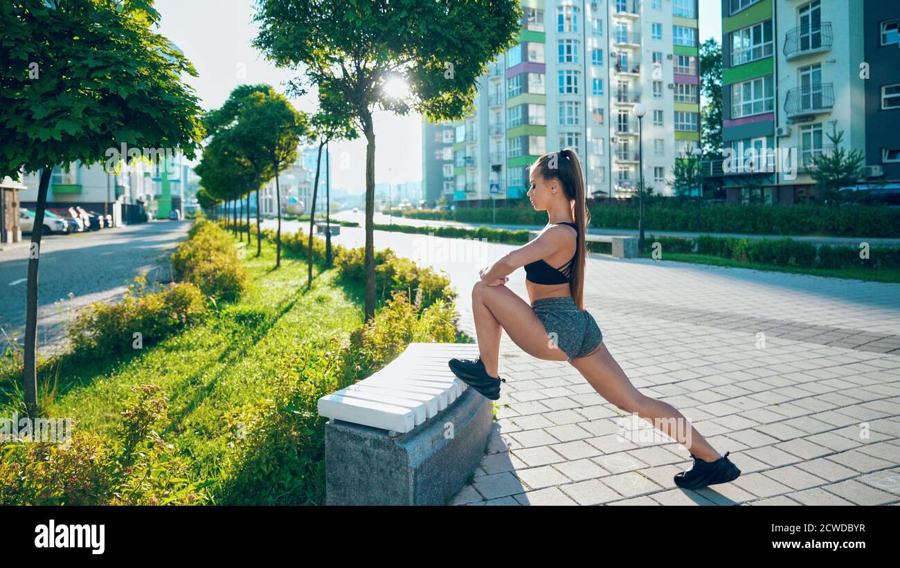 Side view of fitness woman wearing sportswear stretching leg on bench, practicing flexibility exercises after running workout in city streets. Stunning fit woman warming up in rays of morning sun. Stock Photo