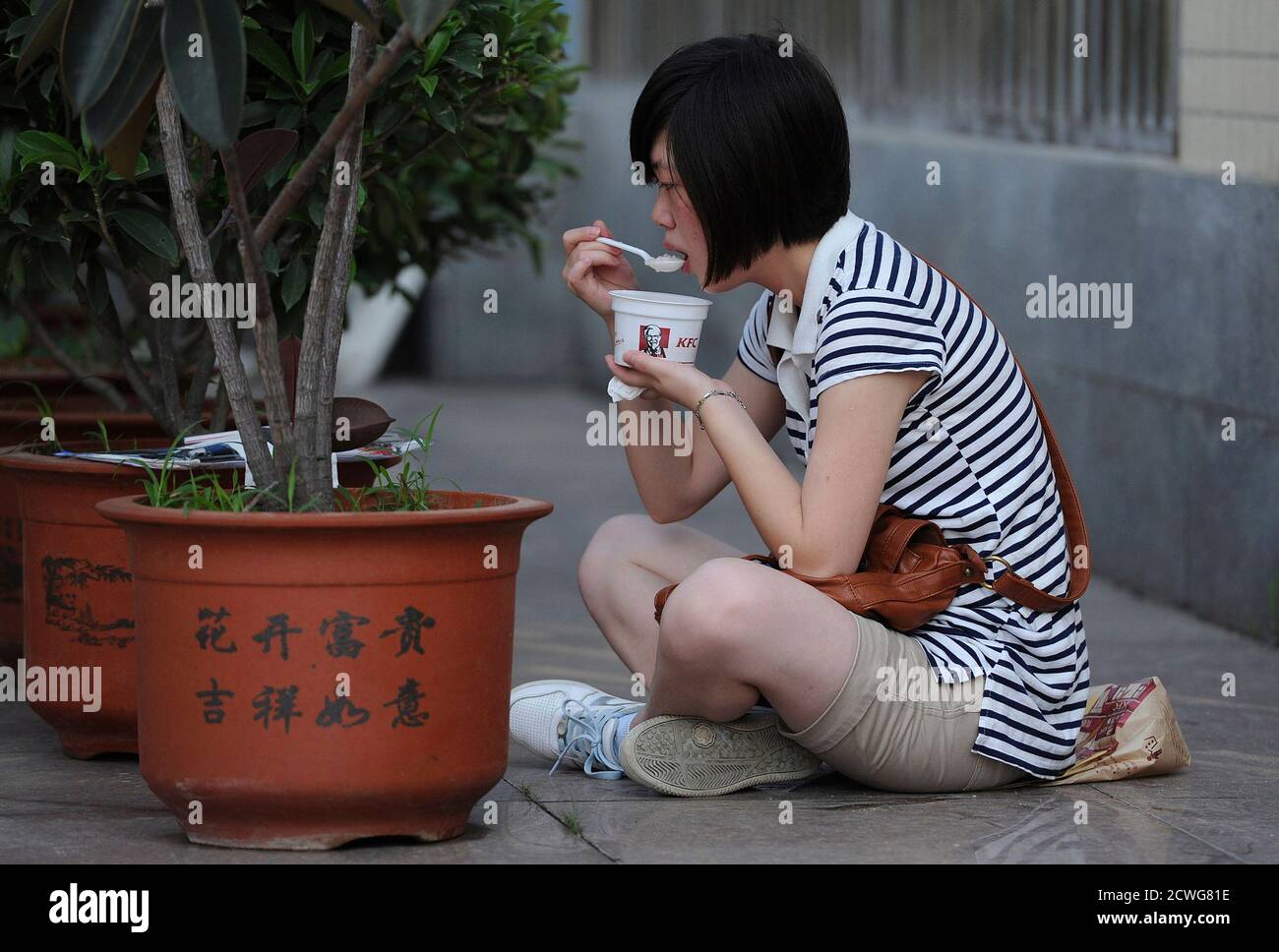 A student sits on the ground as she has a bowl of porridge from Kentucky Fried Chicken (KFC) for breakfast before the second day of National College Entrance Exams in Hefei, Anhui province June 8, 2011. About 9.33 million students make a start on China's national college entrance exams or 'gaokao' from June 7 to 8, a fiercely competitive test that is seen as make-or-break for getting ahead. The Chinese characters on the flowerpot read 'Fortune comes with blooming flowers, good luck and happiness as one wishes'.  REUTERS/Stringer (CHINA - Tags: EDUCATION FOOD ODDLY) Stock Photo