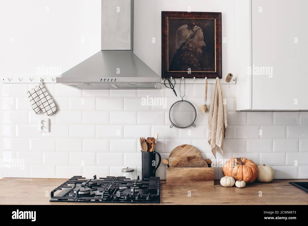 Modern eclectic kitchen interior with pumpkins. White brick wall with metro tiles, peg rails and oil painting. Wooden countertop, hood and gas stove Stock Photo