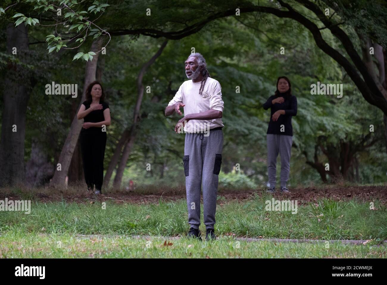A slender older man with long hair leads an exercise class that's a combination of yoga, tai chi & stretching. In a park in Queens, New York City. Stock Photo