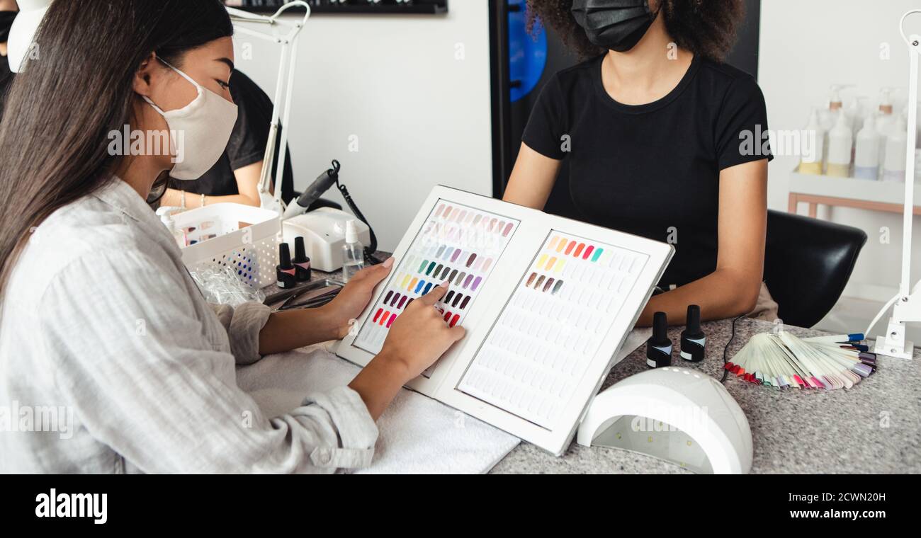 New manicure after lockdown and choice of modern color after quarantine. Asian woman chooses nail polish in catalog with african american master Stock Photo