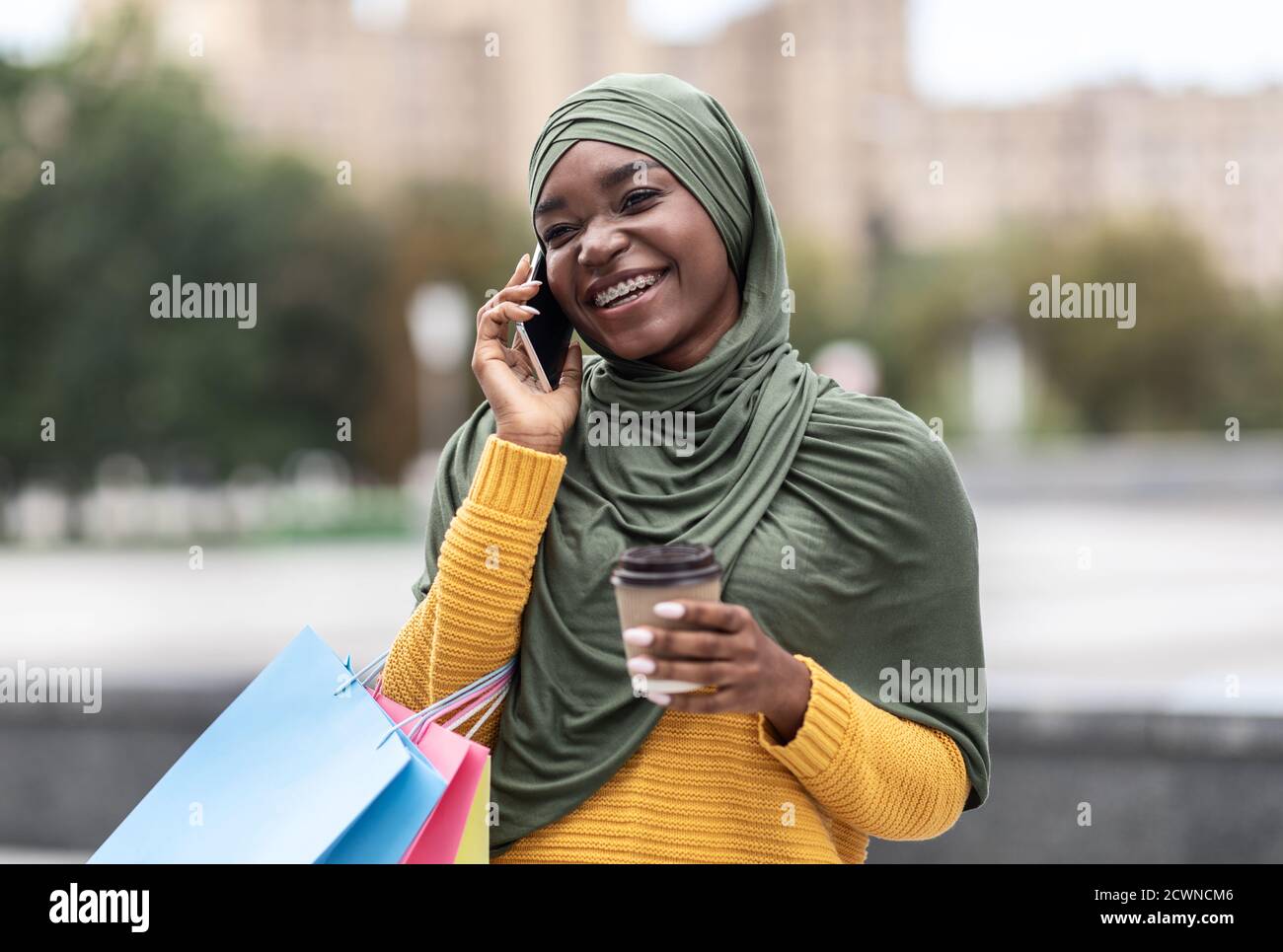 Black Muslim Woman With Shopping Bags And Coffee Talking On Cellphone Outdoors Stock Photo