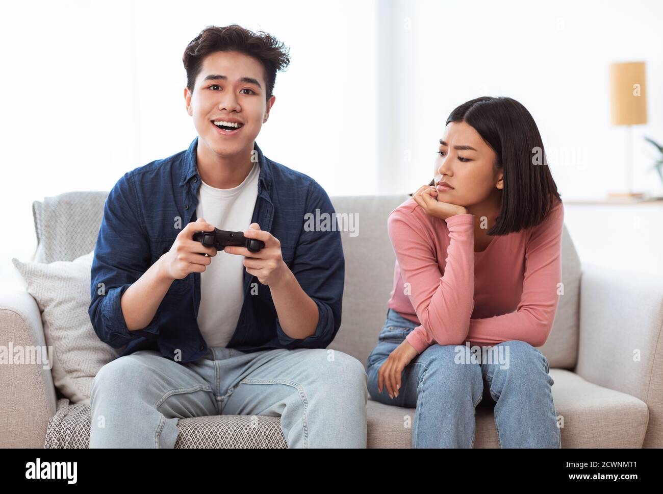 Discontented Asian Wife Looking At Husband Playing Video Game Indoors Stock Photo