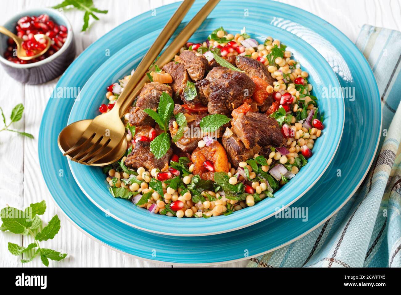 Lamb Stew with Herbed Pearl Couscous and pomegranate seeds in a blue bowl on a white wooden table, horizontal view from above, close-up Stock Photo