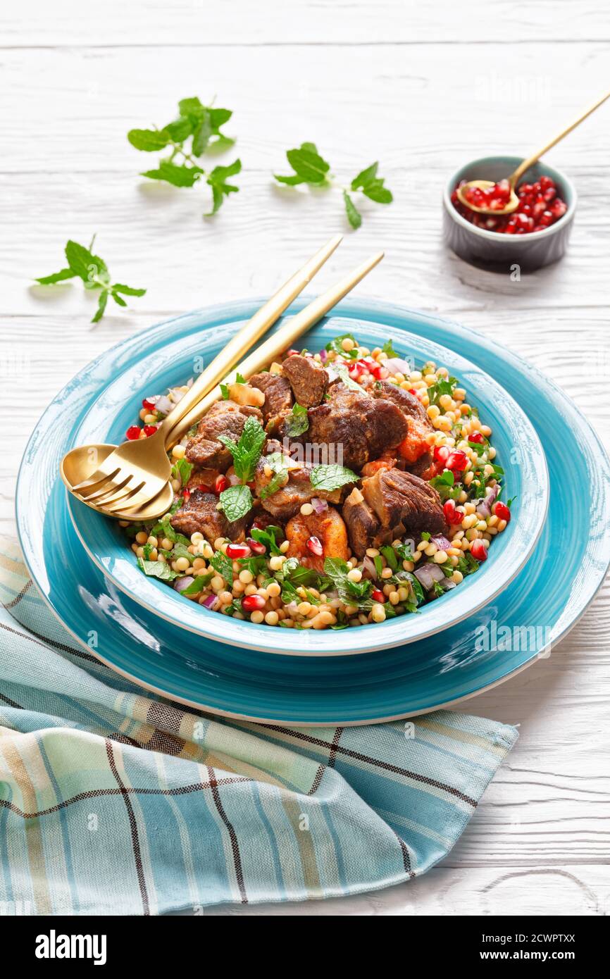 Moroccan Lamb Stew with Herbed Pearl Couscous and pomegranate seeds in a blue bowl on a white wooden table, vertical view from above Stock Photo