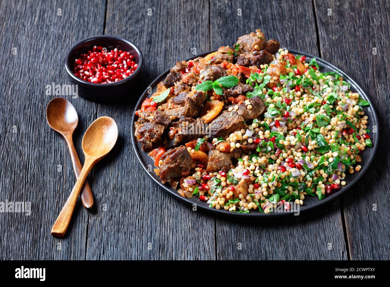 Lamb Stew with Herbed Pearl Couscous and pomegranate seeds on a black plate on a dark wooden table, horizontal view from above Stock Photo