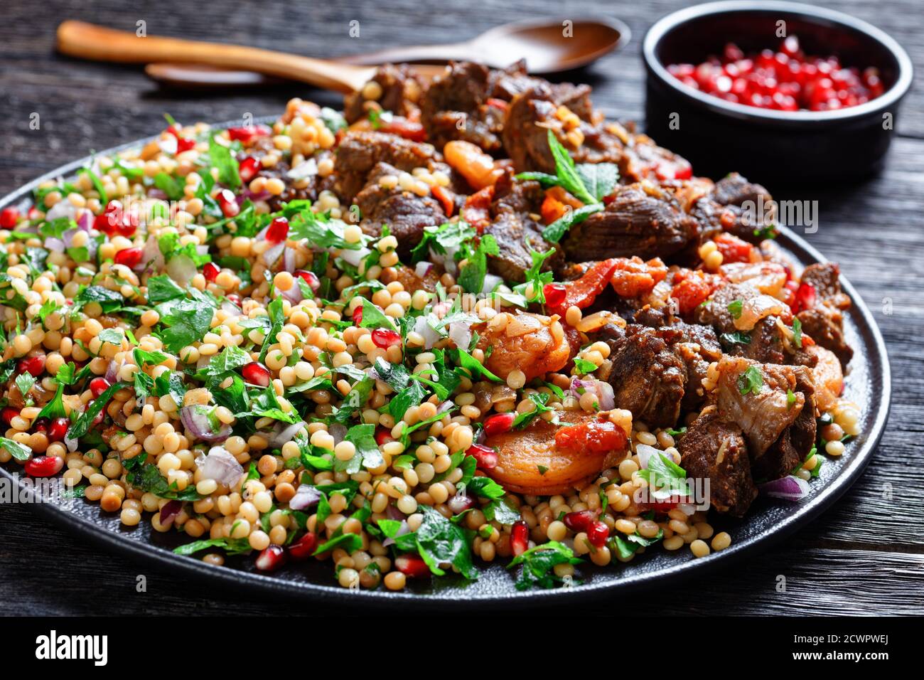 Moroccan Lamb Stew with Herbed Pearl Couscous and pomegranate seeds on a black plate on a dark wooden table, horizontal view from above, close-up Stock Photo